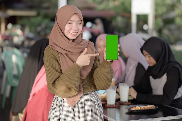 portret van een glimlachend jong Aziatisch tienermeisje dat een leeg groen telefoonscherm toont en wijst.