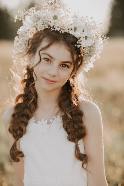 Portret van een glimlachend jong meisje in een witte jurk en een bloemenkroon op haar haar met vlechten op het gebied van de zomer