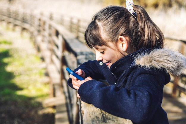 Portret van een glimlachend Kaukasisch meisje dat haar smartphone in het park op een wazig oppervlak gebruikt