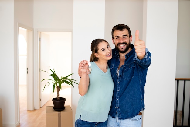 Portret van een glimlachend Kaukasisch paar dat duimen omhoog houdt en de sleutel van hun nieuwe appartement onroerend goed.