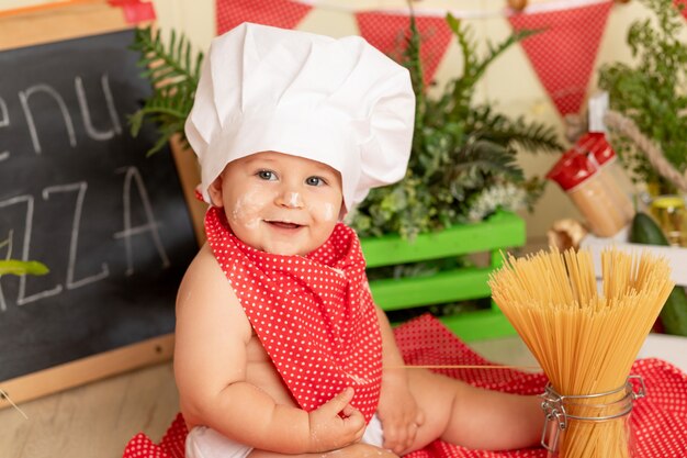 Portret van een glimlachend klein kind in een koksmuts die spaghetti in de keuken voorbereidt