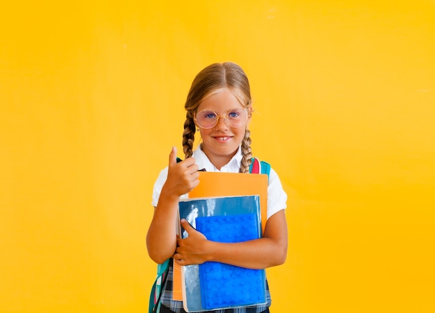 Portret van een glimlachend klein schoolmeisje met rugzak met notitieboekje over gele achtergrond