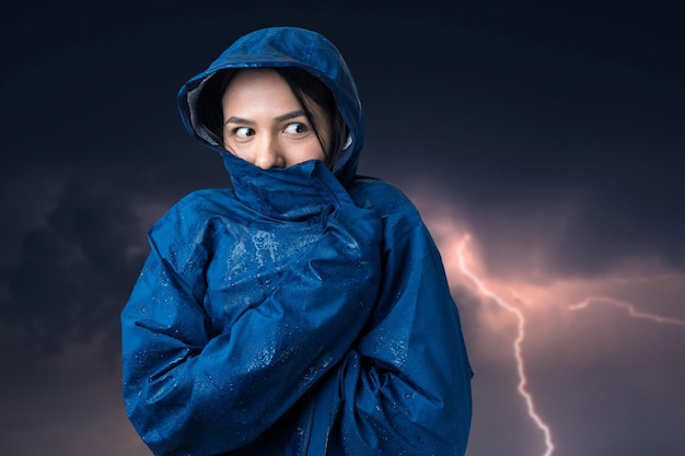 Portret van een glimlachend meisje gekleed in blauwe regenjas in druppels poserend met capuchon tegen de achtergrond van een sombere hemel met bliksem