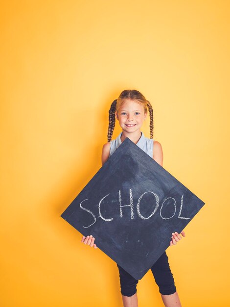 Foto portret van een glimlachend meisje met een bord met schooltekst terwijl het tegen een gele achtergrond staat