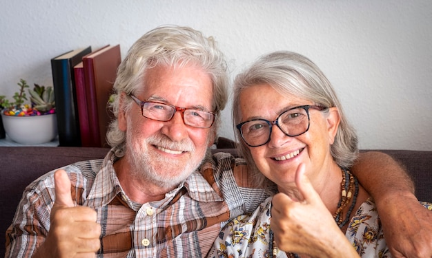 Portret van een glimlachend senior koppel dat thuis op de bank zit en naar de camera kijkt die een positief gebaar doet, wit haar - gelukkig pensioenconcept met twee mensen samen