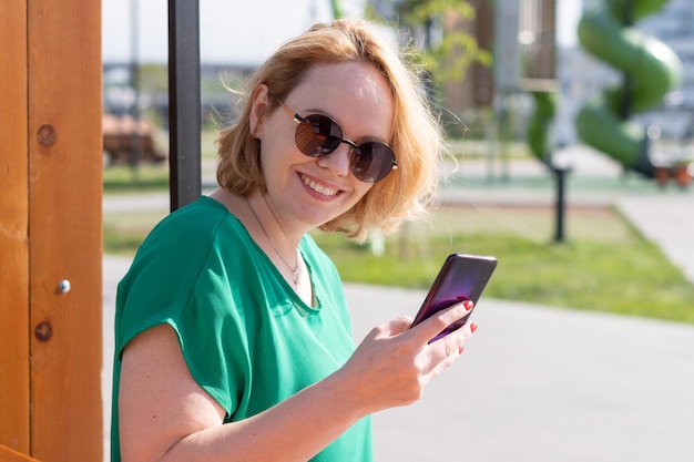 Portret van een glimlachend studentenmeisje in zonnebril met een smartphone in haar handen zittend op een bankje in een stadspark