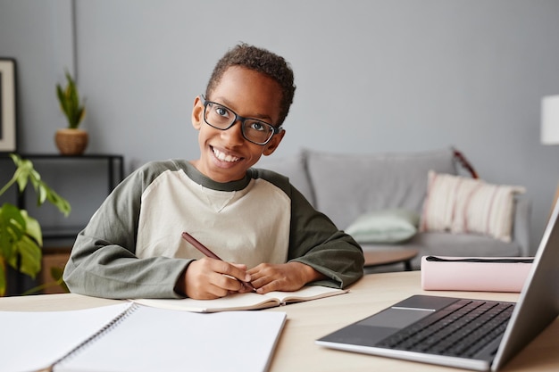 Portret van een glimlachende Afro-Amerikaanse jongen die een bril draagt terwijl hij thuis studeert, homeschooling concept, kopieer ruimte