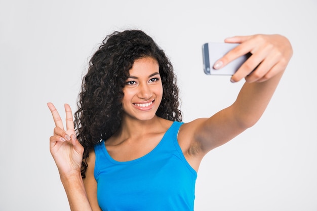 Portret van een glimlachende afro-Amerikaanse vrouw die selfiefoto maakt terwijl twee vingers wordt getoond teken dat op een witte muur wordt geïsoleerd