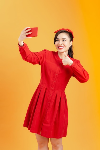 Portret van een glimlachende Aziatische vrouw in rode jurk die selfie foto maakt op smartphone geïsoleerd op een oranje achtergrondxA