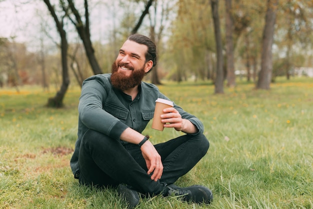 Portret van een glimlachende bebaarde man die in het park op gras zit en een kopje koffie drinkt