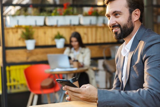 Portret van een glimlachende bebaarde man met een jas die afhaalkoffie drinkt en een mobiele telefoon gebruikt terwijl hij in een café zit