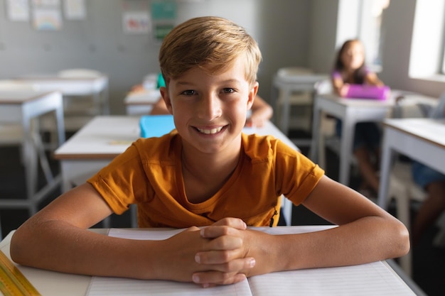 Portret van een glimlachende blanke basisschooljongen met gebonden handen die aan een bureau in de klas zit. ongewijzigd, onderwijs, leren, jeugd, studeren en schoolconcept.