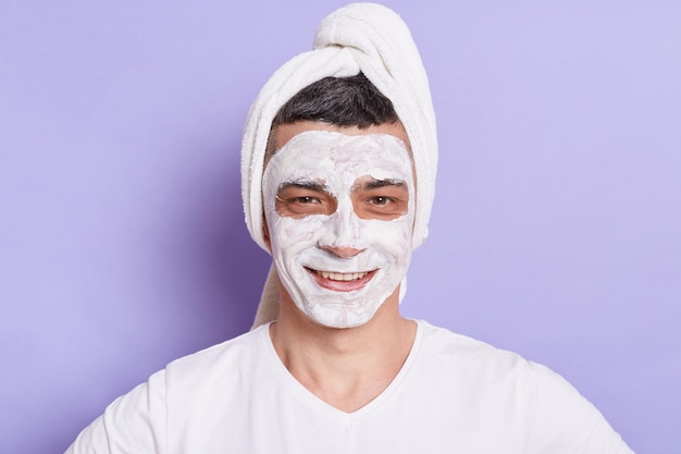 Portret van een glimlachende blanke man met een masker voor de huid gewikkeld in een handdoek geïsoleerd op een violette achtergrond die thuis cosmetische procedures doet
