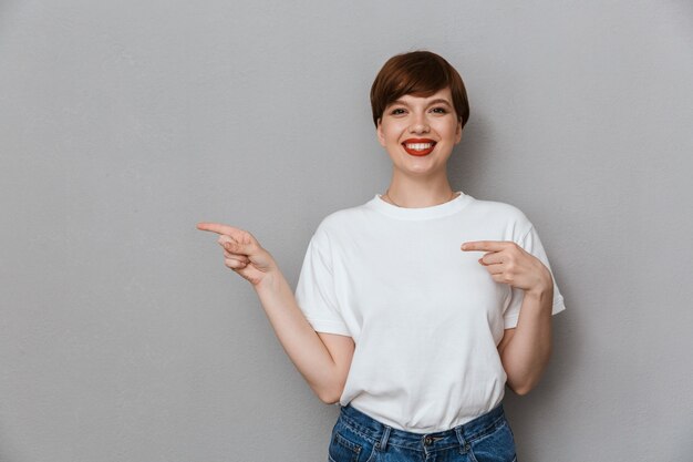 Portret van een glimlachende brunette vrouw met een casual t-shirt die met de vingers opzij wijst naar copyspace geïsoleerd over grijze muur