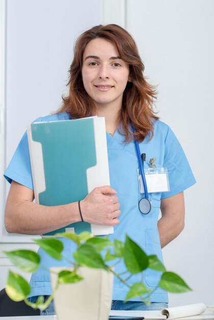 Foto portret van een glimlachende dokter die een dossier vasthoudt terwijl hij tegen een muur in het ziekenhuis staat