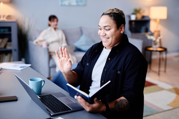 Portret van een glimlachende getatoeëerde vrouw die naar het scherm van de laptop zwaait tijdens online vergaderruimte