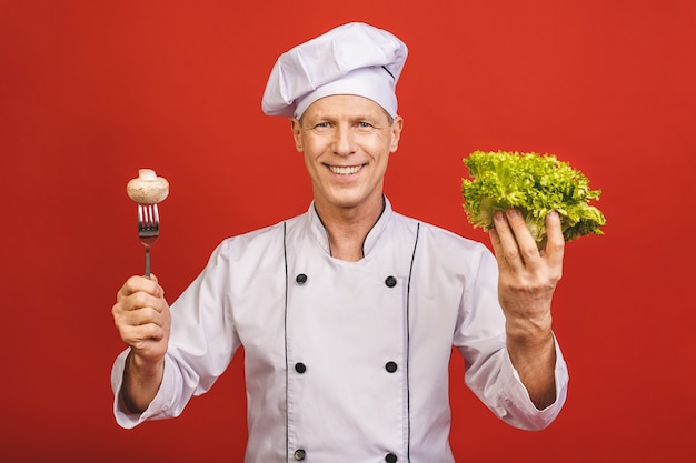 Portret van een glimlachende hogere die de holdingssalade en paddestoel van de chef-kokkok op een rode achtergrond wordt geïsoleerd.