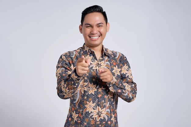 Portret van een glimlachende jonge Aziatische man in een batik shirt die naar de camera kijkt en met de wijsvinger naar u wijst, geïsoleerd op een witte achtergrond
