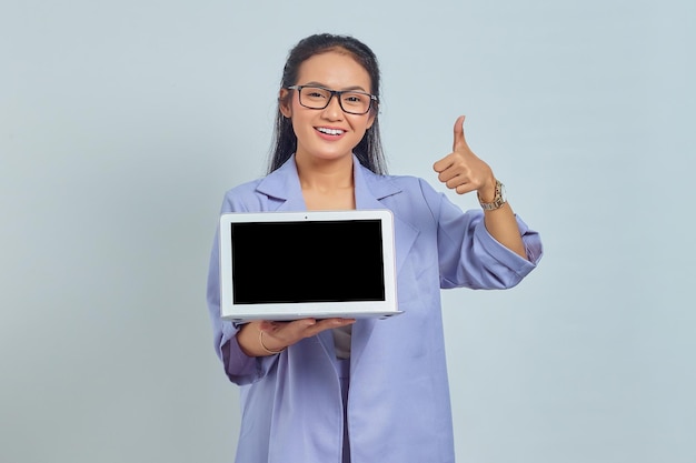 Portret van een glimlachende jonge Aziatische vrouw die een leeg scherm van een laptop toont en een duim omhoog gebaar maakt dat op een witte achtergrond wordt geïsoleerd