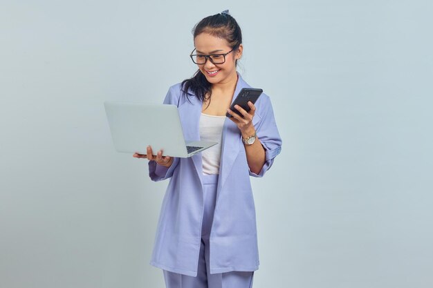 Portret van een glimlachende jonge Aziatische vrouw die naar een laptop kijkt en een mobiele telefoon vasthoudt die op een witte achtergrond wordt geïsoleerd