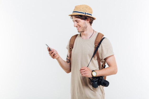 Portret van een glimlachende jonge man met rugzak en camera met behulp van slimme telefoon geïsoleerd op een witte achtergrond