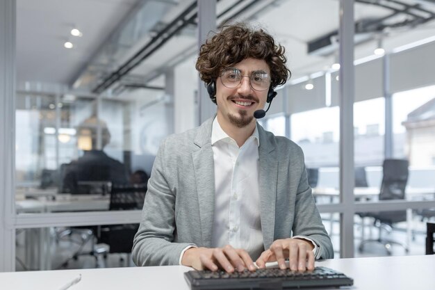 Portret van een glimlachende jonge succesvolle man die een headset draagt en in het kantoor aan een computer werkt