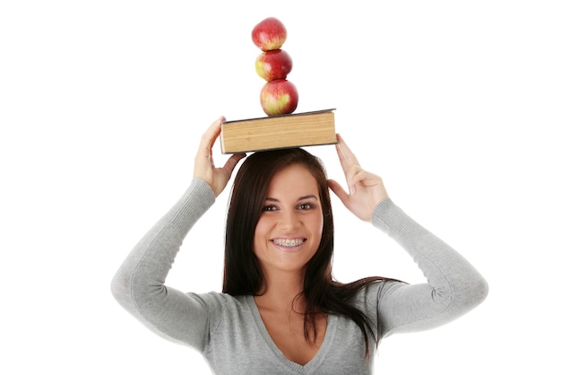 Foto portret van een glimlachende jonge vrouw die appels op een boek over haar hoofd balanceert tegen een witte achtergrond