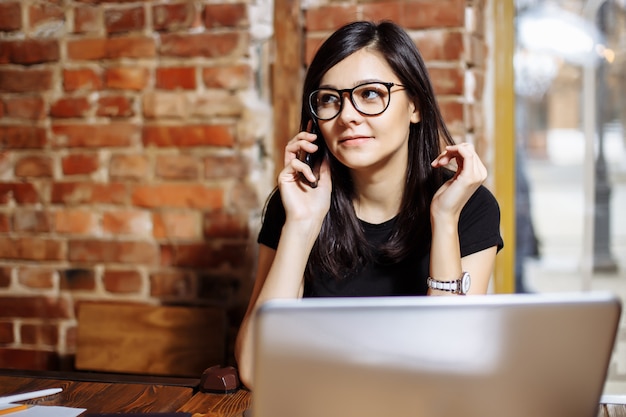 Portret van een glimlachende jonge vrouw die laptop met behulp van en op mobiele telefoon spreekt.