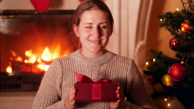 Portret van een glimlachende jonge vrouw die op kerstavond in de woonkamer zit en een cadeau van de kerstman in de camera laat zien