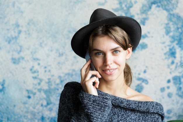 Portret van een glimlachende jonge vrouw gekleed in hoed en trui close-up praten op mobiele telefoon over blauwe muur