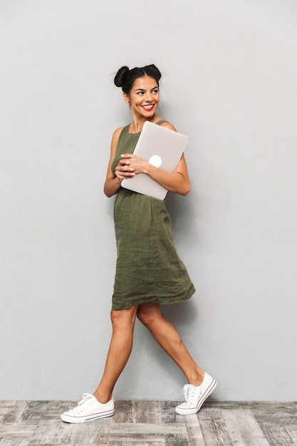 Portret van een glimlachende jonge vrouw in geïsoleerde kleding, die laptop draagt, het lopen