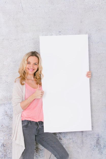 Portret van een glimlachende jonge vrouw met een leeg bord met duimen omhoog die tegen de muur staat