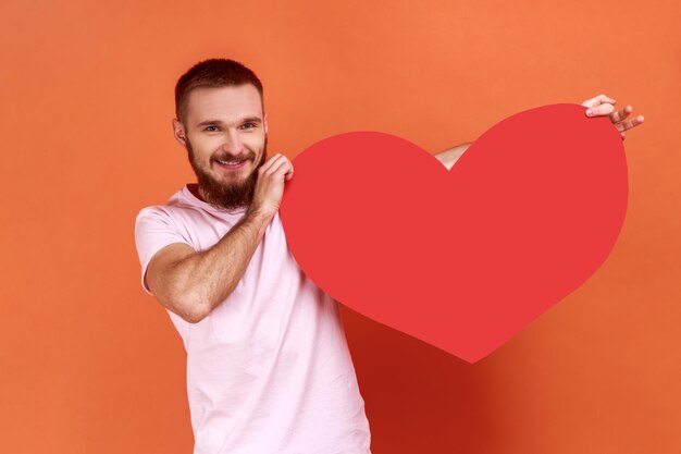 Portret van een glimlachende knappe bebaarde man die een groot rood hart vasthoudt, positieve romantische emoties uitdrukt, een roze T-shirt draagt. Indoor studio opname geïsoleerd op een oranje achtergrond.