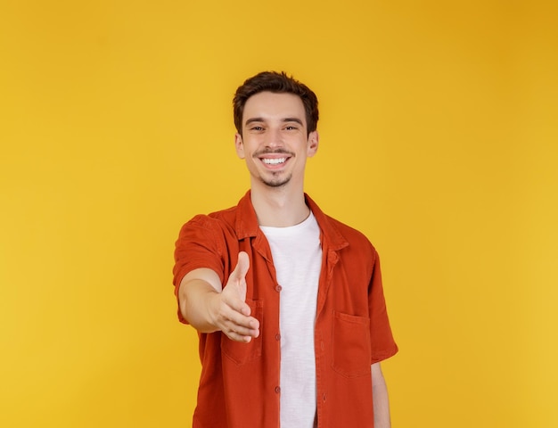 Portret van een glimlachende knappe man die zijn hand uitsteekt voor een handdruk, kijk vriendelijk groet je hallo gebaar over een gele achtergrond