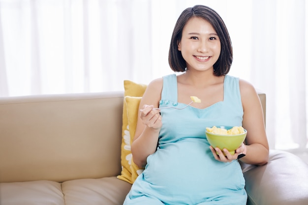 Portret van een glimlachende mooie jonge zwangere vrouw die een kom met gesneden fruit eet voor de lunch