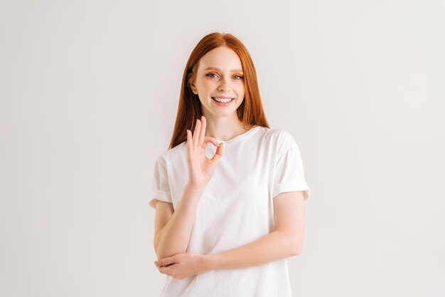 Portret van een glimlachende mooie roodharige jonge vrouw die een goed gebaar toont en naar de camera kijkt, staande op een witte geïsoleerde achtergrond in de studio. fronvmening van vrolijke aantrekkelijke vrouw die ok tekengebaar toont