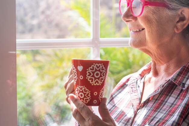 Portret van een glimlachende senior vrouw met een roze bril die bij het raam staat en door een oudere, grijze, harige, blanke grootmoeder kijkt die een rode kop koffie of thee vasthoudt