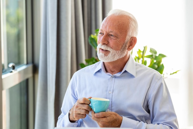 Portret van een glimlachende senior zakenman die een koffiemok vasthoudt en door het raam kijkt, close-up van vrije ruimte