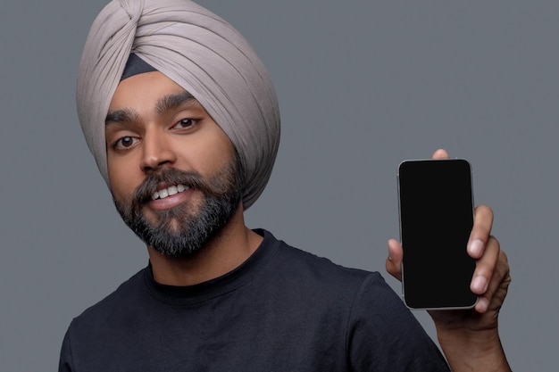 Portret van een glimlachende tevreden man in de tulband die zijn smartphone voor de camera houdt