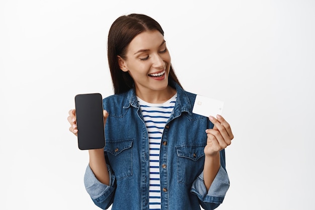 Portret van een glimlachende tevreden vrouw die een creditcard en een mobiele telefoonscherminterface van de speciale verkoop van de online shopping-app op een smartphone toont die een witte achtergrond weergeeft