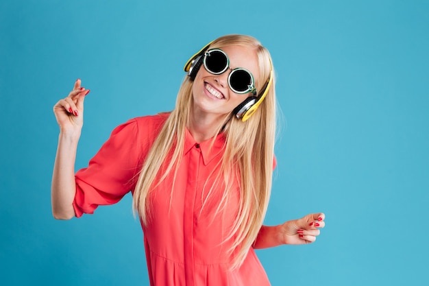 Portret van een glimlachende vrolijke blonde vrouw in zonnebril die muziek luistert met een koptelefoon en danst over blauwe muur