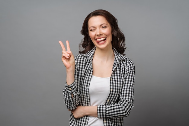 Portret van een glimlachende vrouw die een overwinningsteken toont en een camera bekijkt die op grijs wordt geïsoleerd