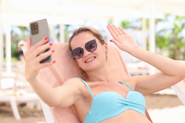 Portret van een glimlachende vrouw die op een ligstoel op het strand ligt en met haar hand zwaait in smartphone