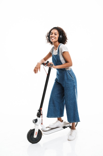 Portret van een glimlachende vrouw gekleed in een denim overall met een koptelefoon tijdens het rijden op een scooter geïsoleerd over een witte muur