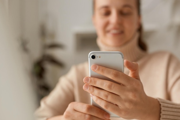 Portret van een glimlachende vrouw met een wazig gezicht met een beige trui die een smartphone gebruikt die nieuws leest of berichten typt die positieve emoties uitdrukken