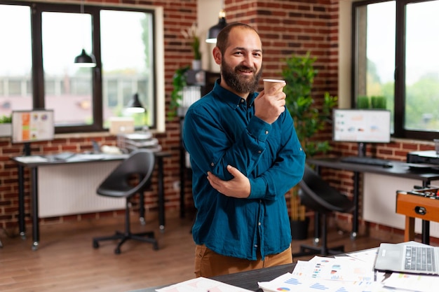 Portret van een glimlachende zakenman die een kopje koffie vasthoudt en in het kantoor van een opstartend bedrijf staat en bedrijfsdocumenten analyseert die werken bij een marketingpresentatie. Uitvoerend manager planning financiële strategie