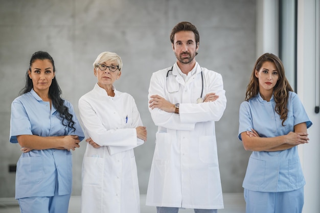 Portret van een glimlachende zelfverzekerde arts die met gekruiste armen in een ziekenhuisgang staat met zijn collega's op de achtergrond.