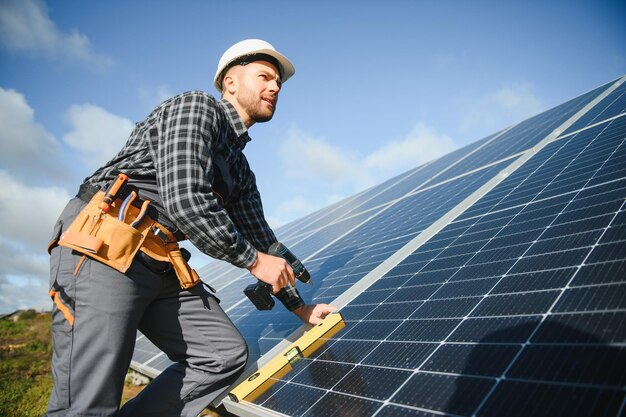 Portret van een glimlachende zelfverzekerde ingenieur-technicus met een elektrische schroevendraaier die voor een onafgewerkt hoog fotovoltaïsch zonnepaneel staat