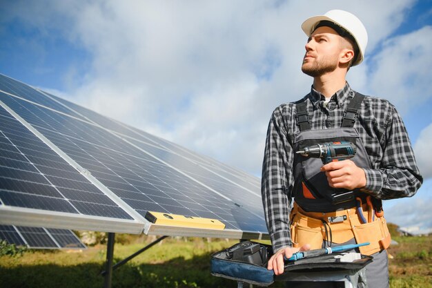 Portret van een glimlachende zelfverzekerde ingenieur-technicus met een elektrische schroevendraaier die voor een onafgewerkt hoog fotovoltaïsch zonnepaneel staat