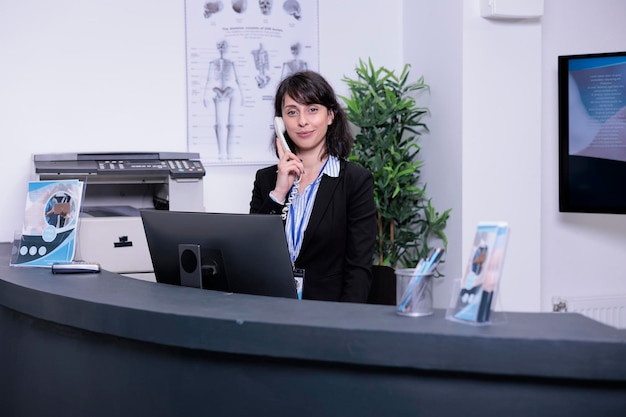 Portret van een glimlachende ziekenhuisreceptionist die de oproep van de patiënt beantwoordt om een afspraak te maken voor klinisch consult in een privékliniek. Professioneel ogende vrouw die werkt bij de receptie met behulp van de telefoon.